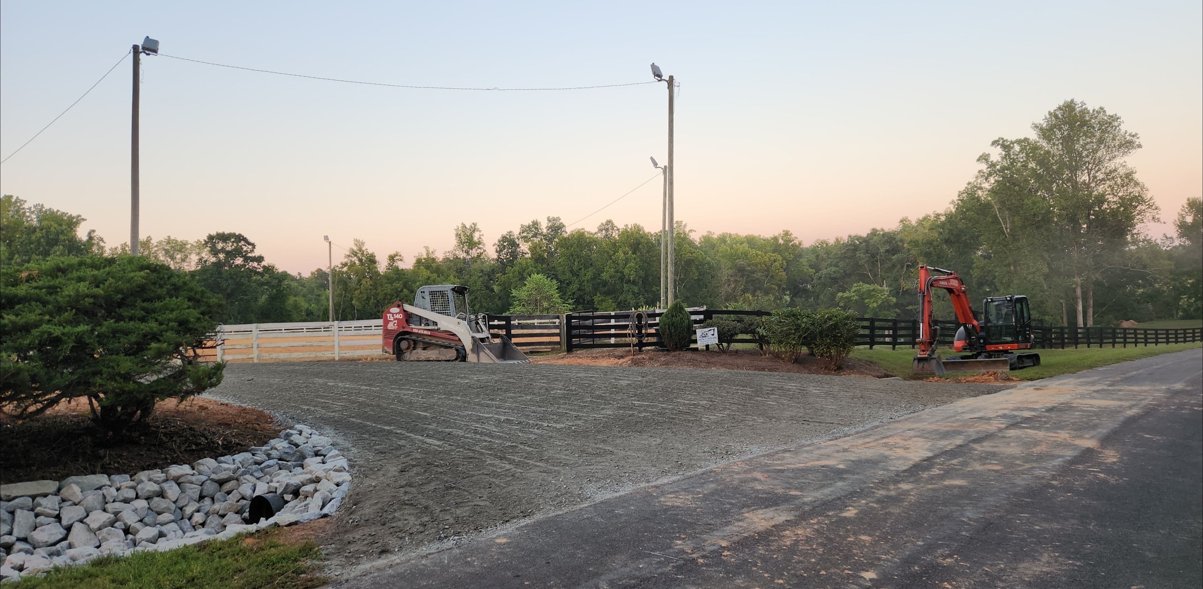 Construction equipment on a gravel site near a horse arena at sunrise.