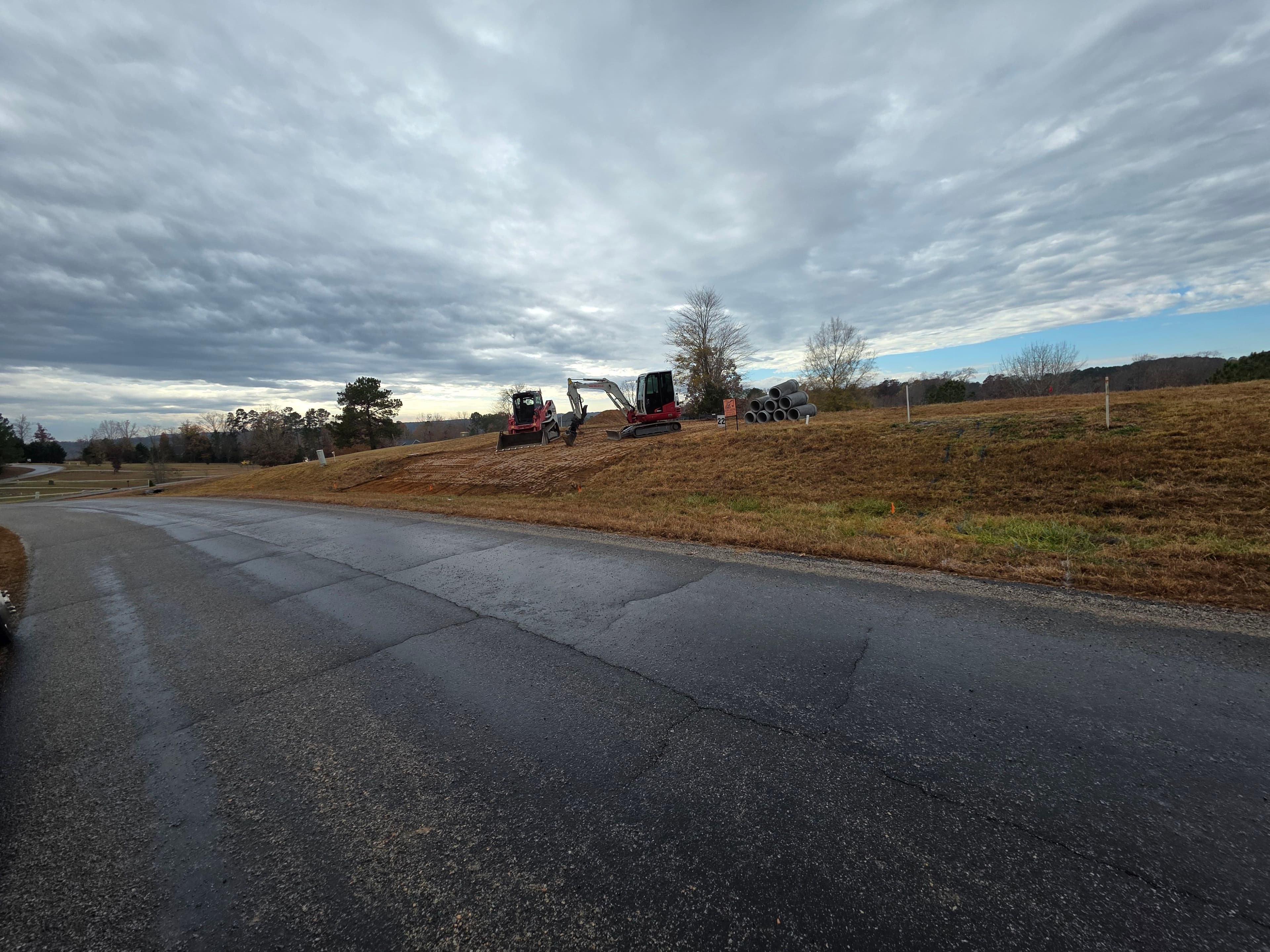 Construction equipment on a grassy hill by a road under a cloudy sky.