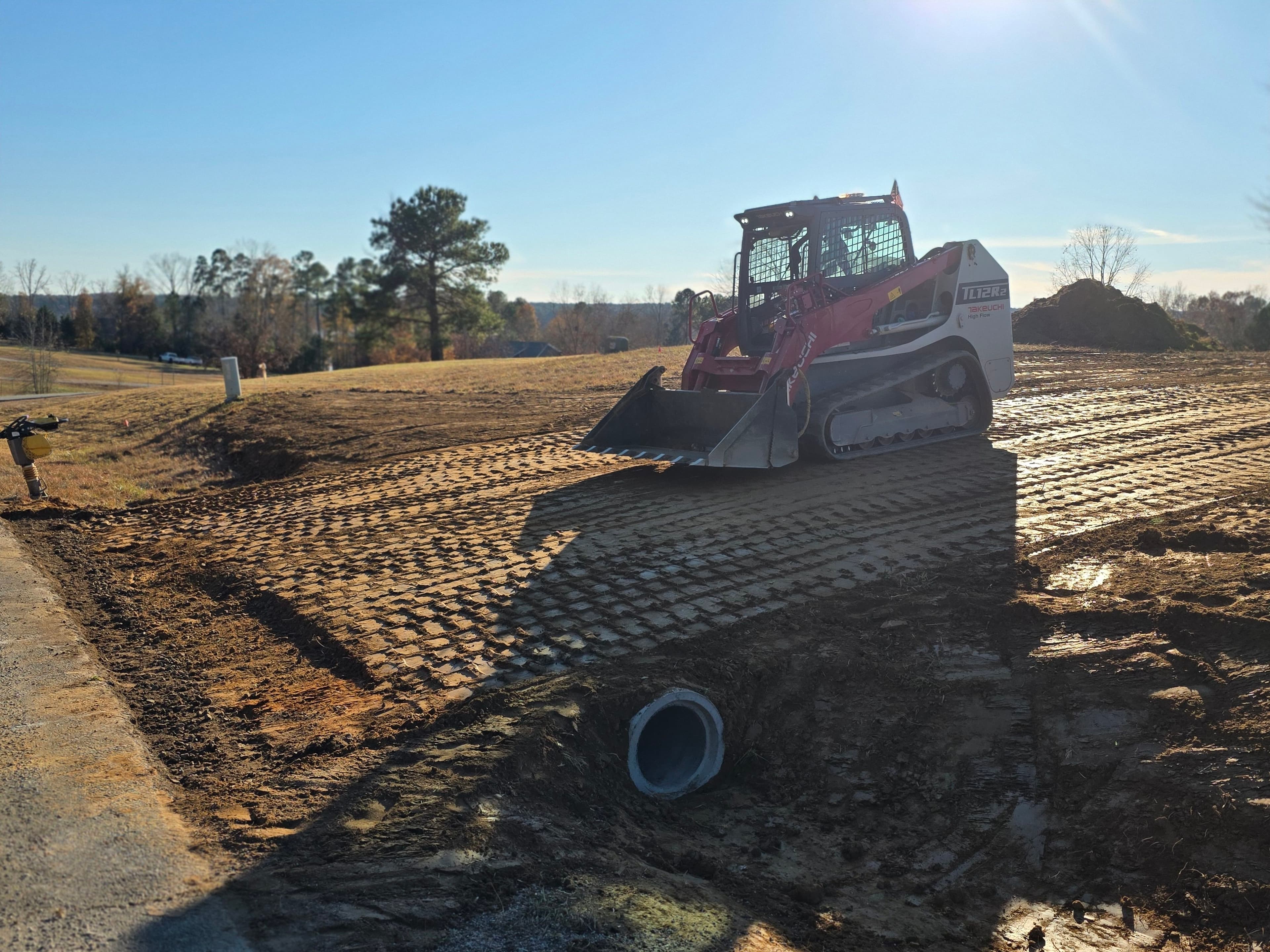 Skid steer loader on a construction site preparing ground with tire tracks and drainage pipe.