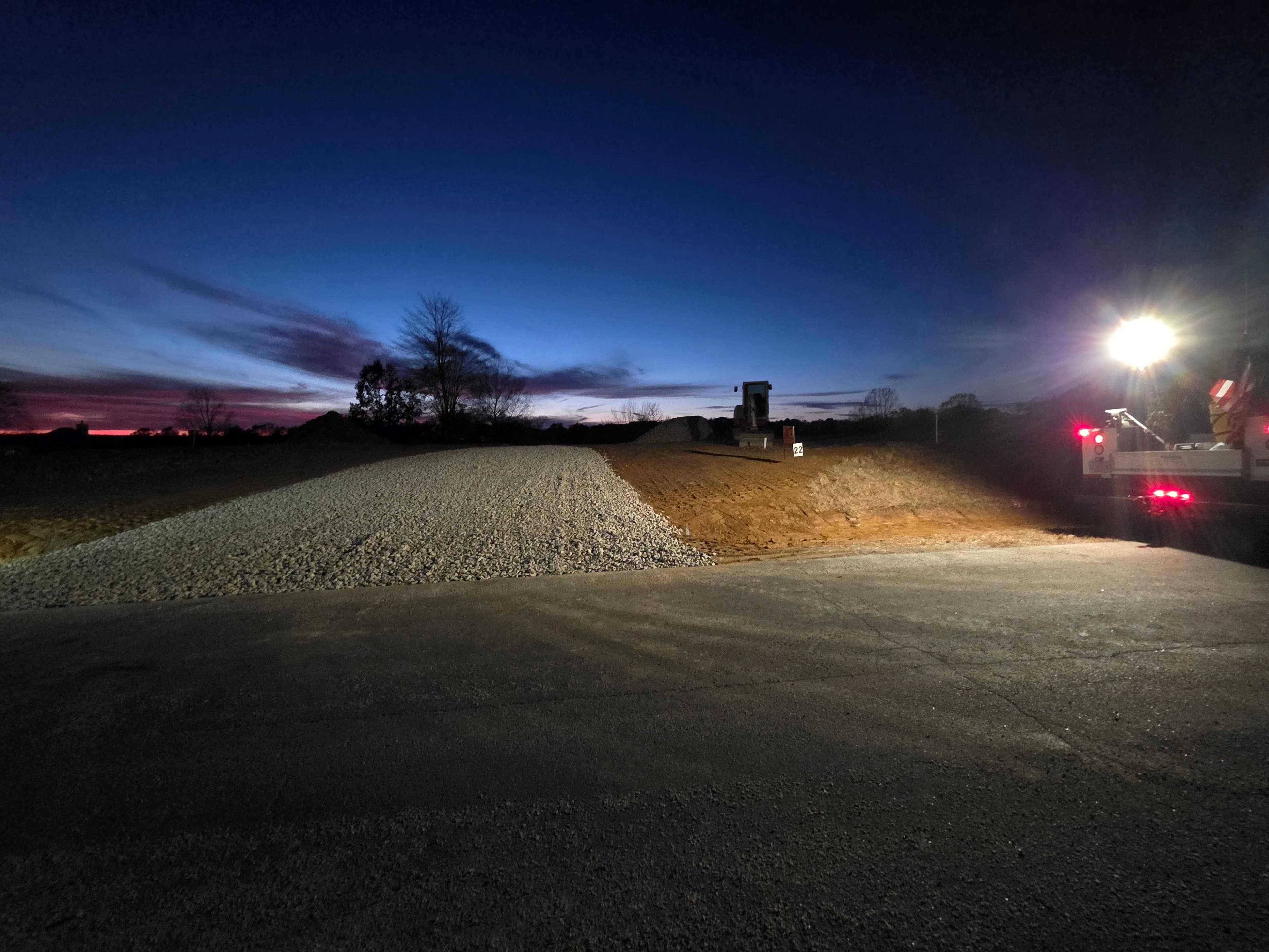 Construction site at dusk, featuring gravel and illuminated machinery against a twilight sky.