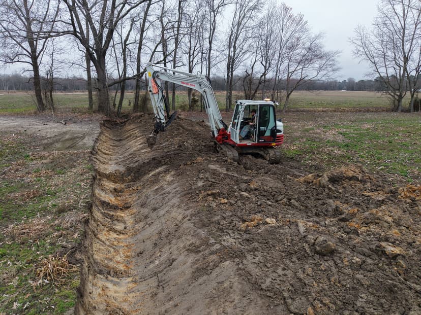 Excavator digging a trench in a rural area surrounded by trees and bare ground.
