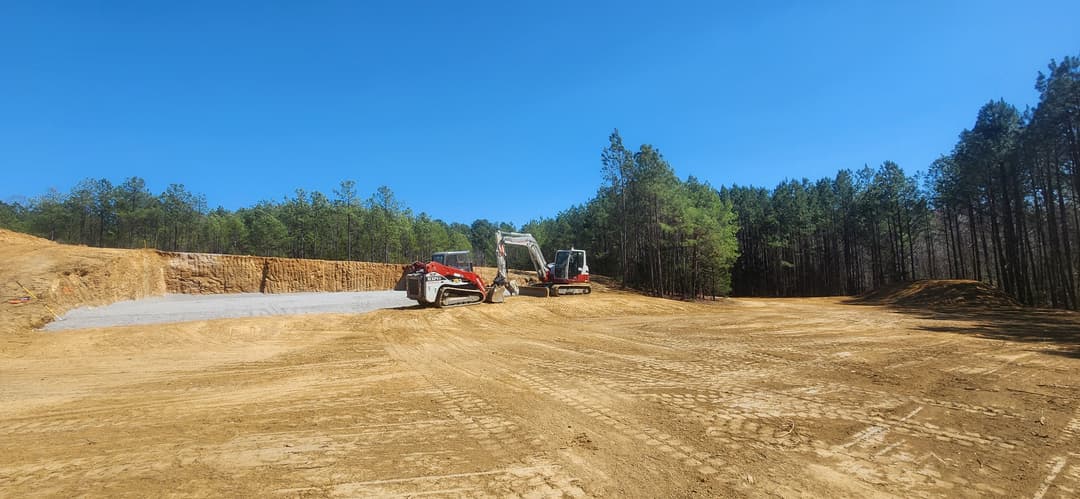 Construction site with heavy machinery on a clear day, surrounded by pine trees.