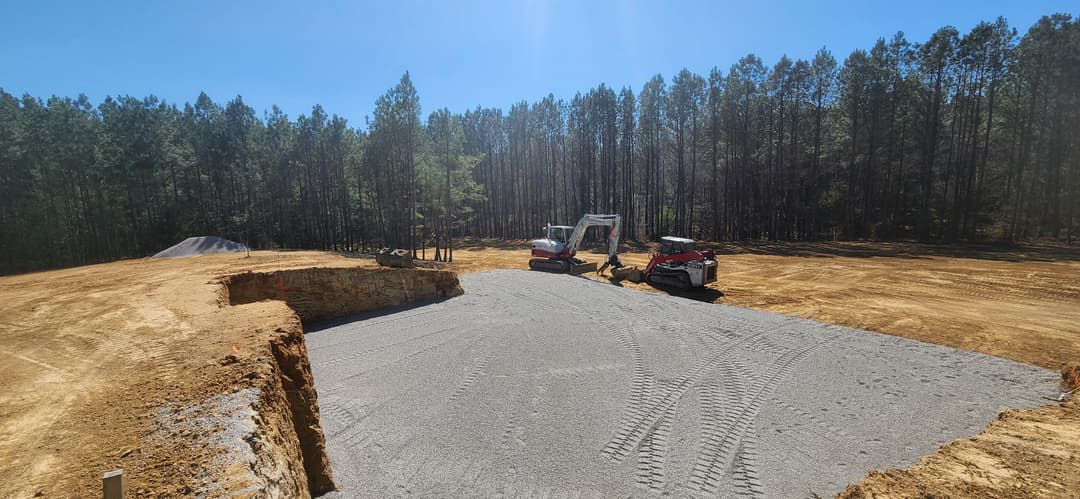 Construction site with heavy machinery preparing a gravel foundation in a forested area.