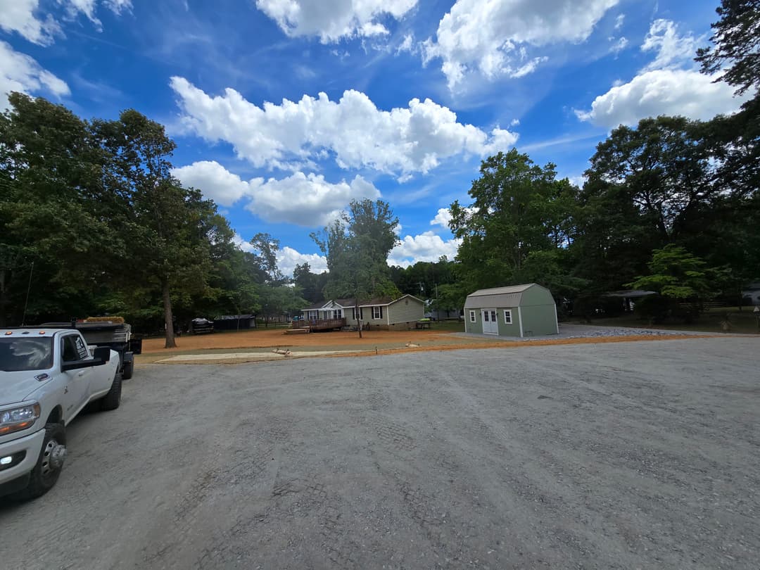 Gravel area with a truck, house, and shed under a blue sky with clouds.