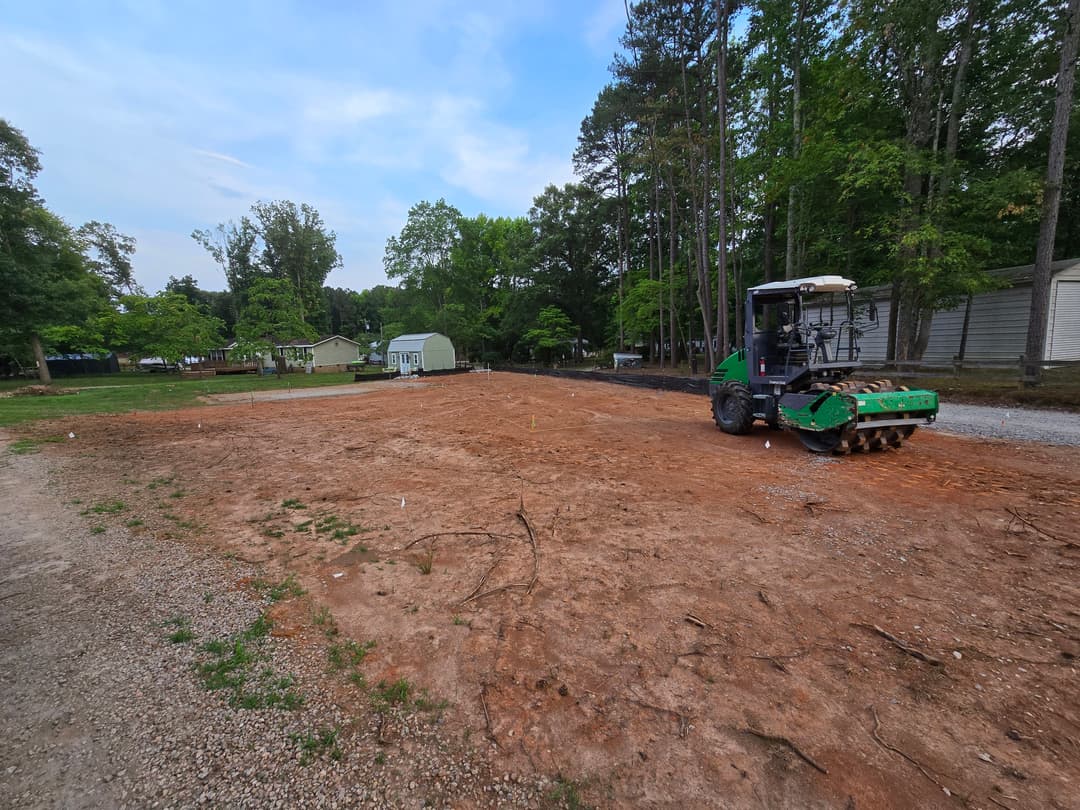 Construction equipment leveling a cleared lot surrounded by trees and nearby buildings.