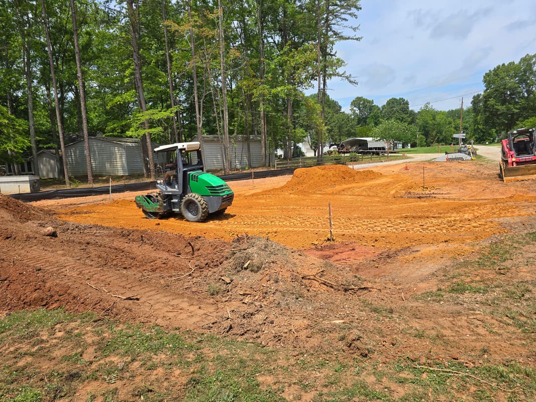 Construction site with machinery moving dirt among trees and gravel road under a clear blue sky.