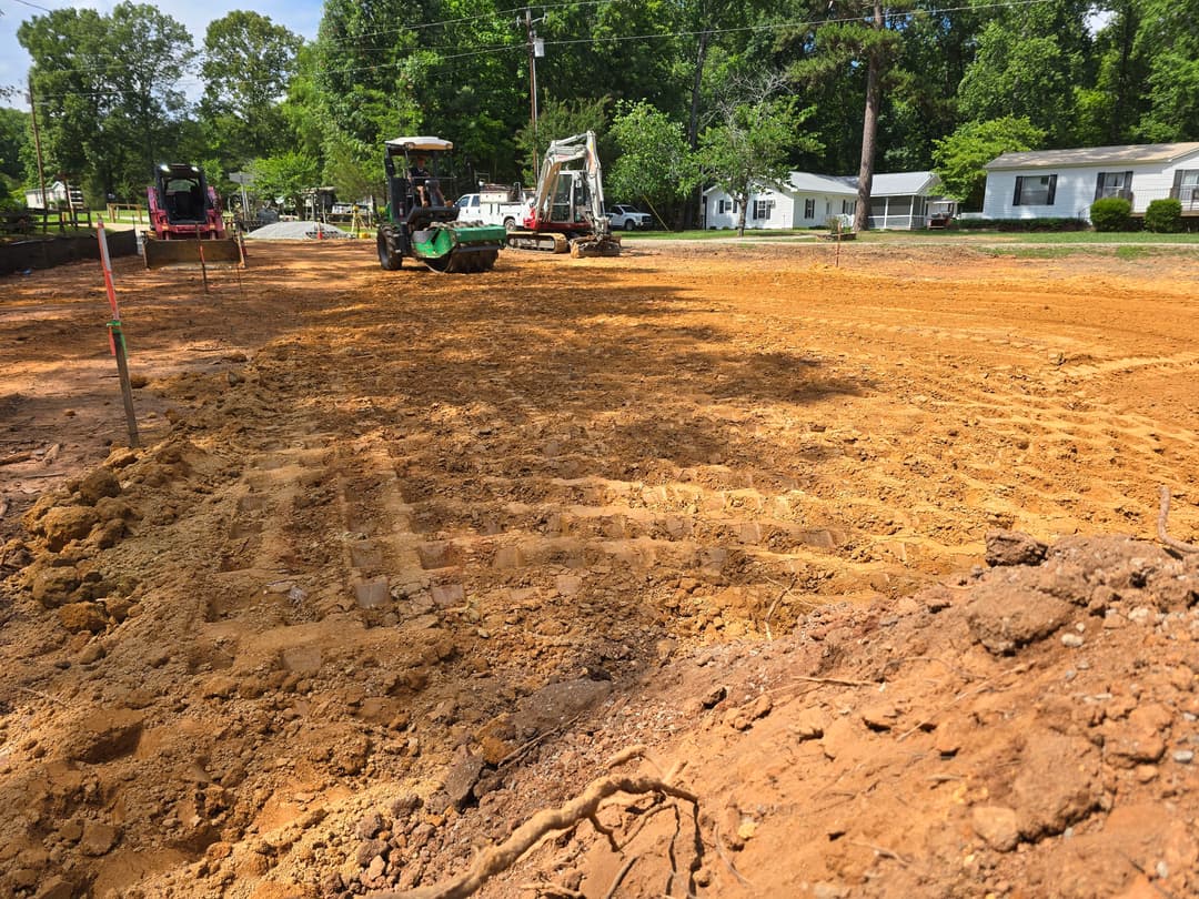 Construction site with machinery on dirt road, surrounded by trees and residential homes.