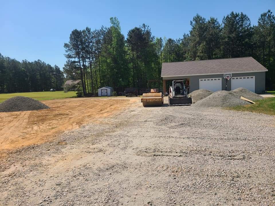 Construction site with gravel piles and heavy machinery near a garage in a wooded area.