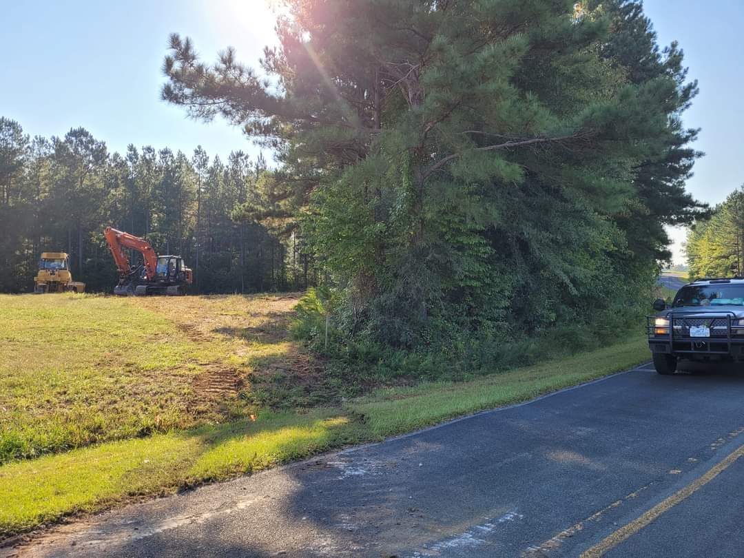 Excavators working on a clearing site beside a roadside with police vehicle nearby.