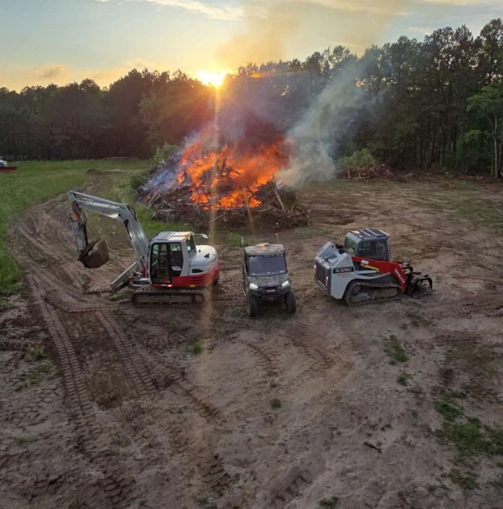 Excavators and tractors near a large bonfire at sunset in a rural landscape.