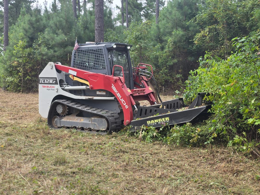 Skid steer loader clearing brush in a wooded area, showcasing heavy-duty equipment in use.