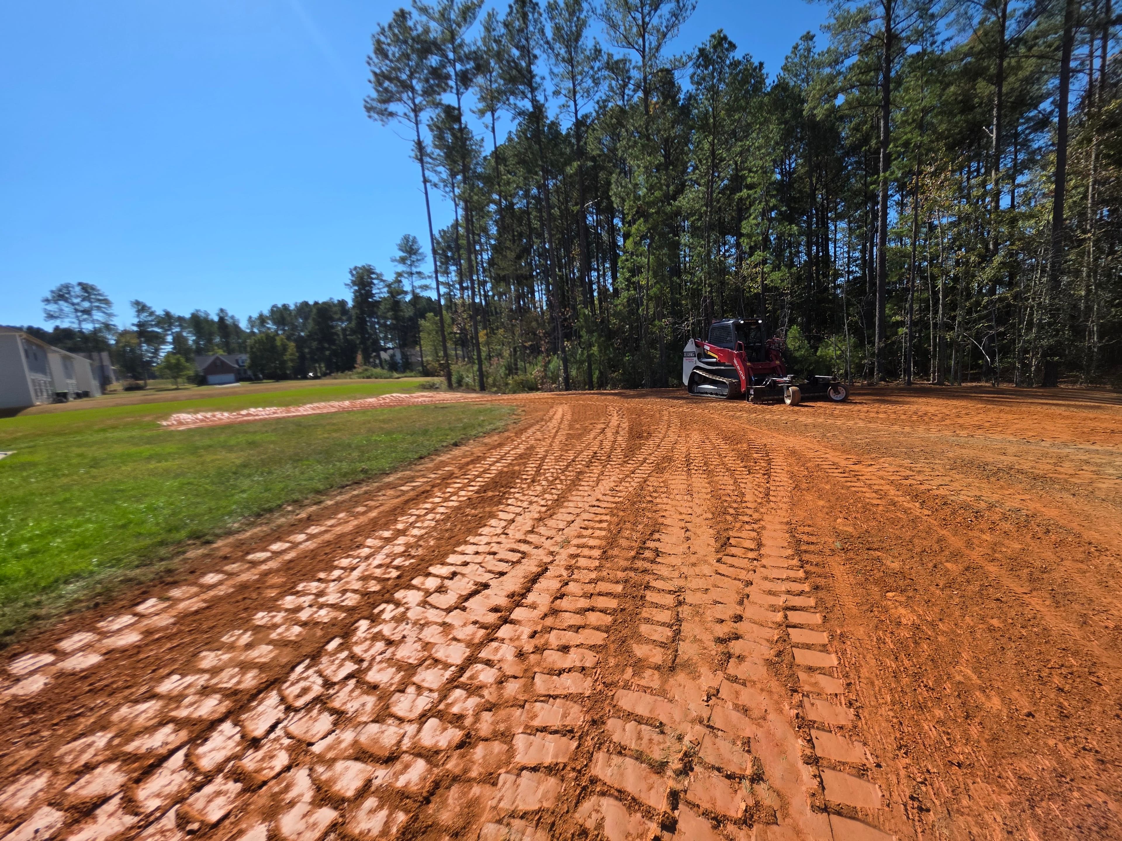 Tractor on freshly tilled red dirt lot surrounded by trees under a clear blue sky.