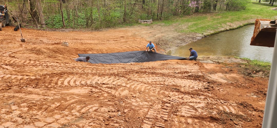 Workers installing a black tarp on a dirt area near a pond, preparing for landscaping.