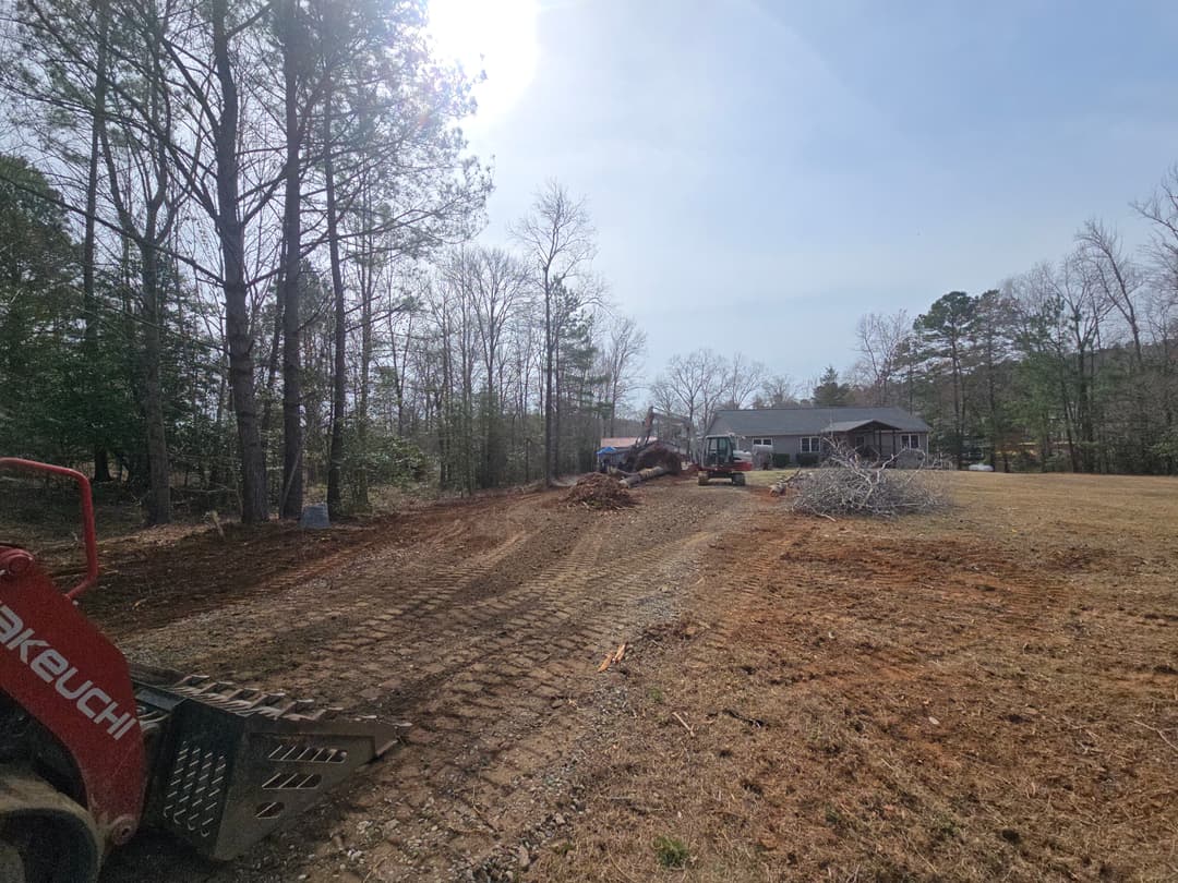 Excavator clearing land near a house with fallen branches in a wooded area.