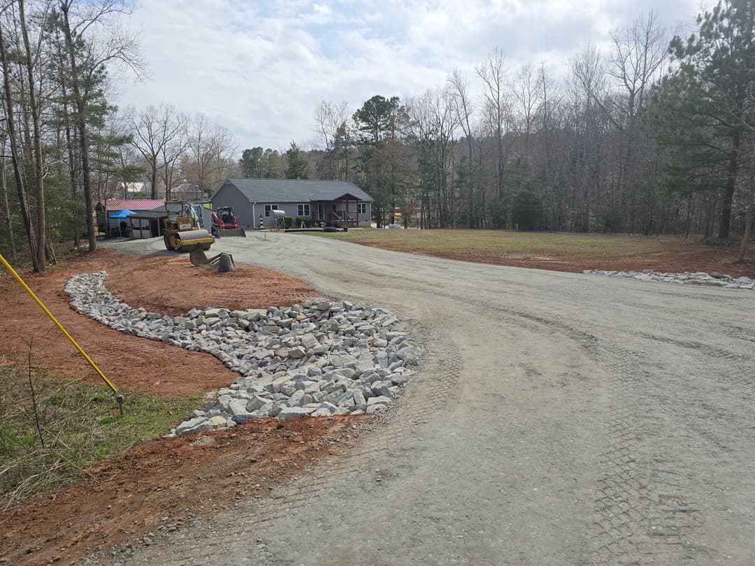 Gravel driveway and landscape featuring rocks with a house and trees in the background.