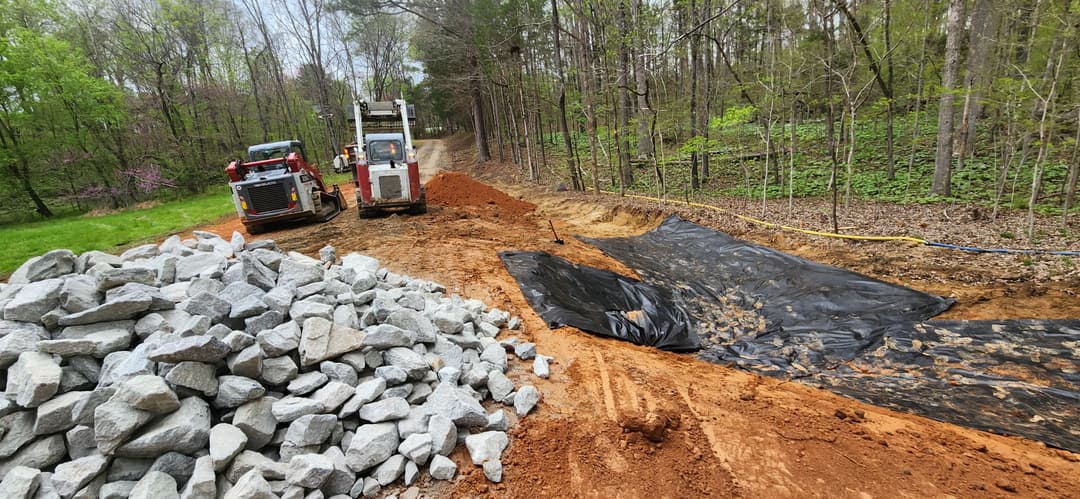 Construction site with heavy machinery, gravel pile, and erosion control fabric along dirt path.