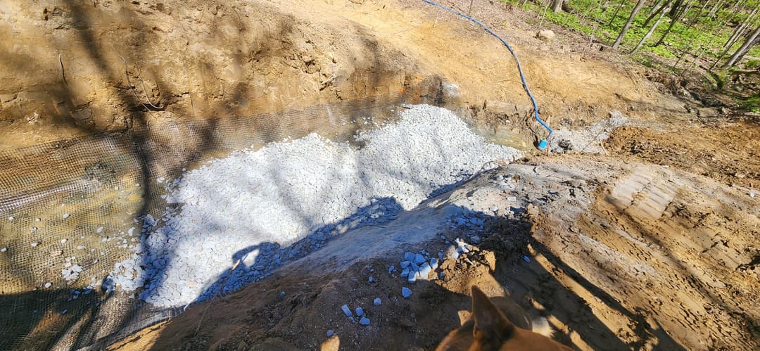 Excavated pit filled with gravel, surrounded by mesh, in a wooded area.