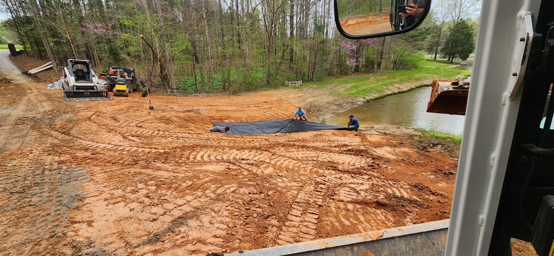 Construction workers laying protective fabric near a waterway, with machinery in the background.