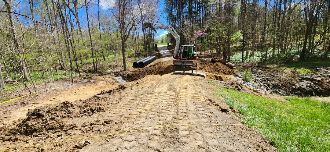 Excavator working on a construction site with newly laid pipe in wooded area.