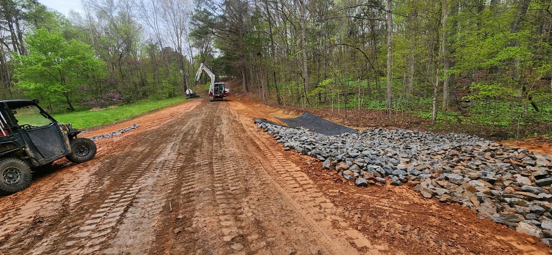 Construction site showing gravel and machinery on a dirt road in a wooded area.