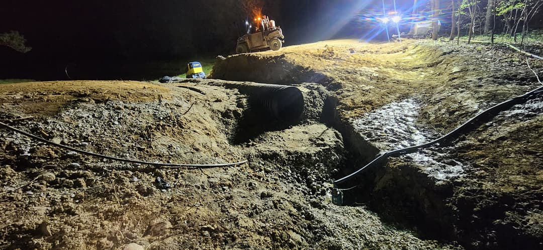 Construction site at night with heavy machinery, excavated ground, and lighting setup.