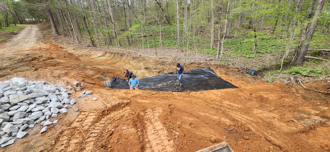 Workers laying protective fabric on a construction site amid trees and gravel.