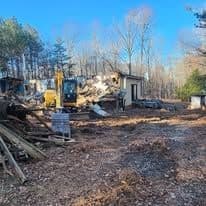 Demolished house with construction machinery on a wooded lot, debris scattered around.