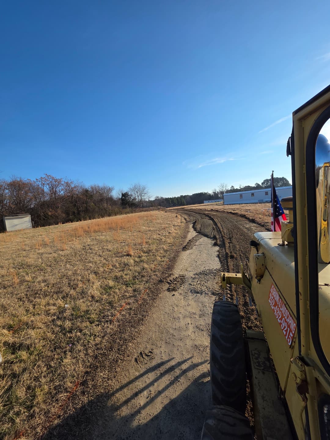 Excavator clearing a dirt road with an American flag, blue sky in the background.
