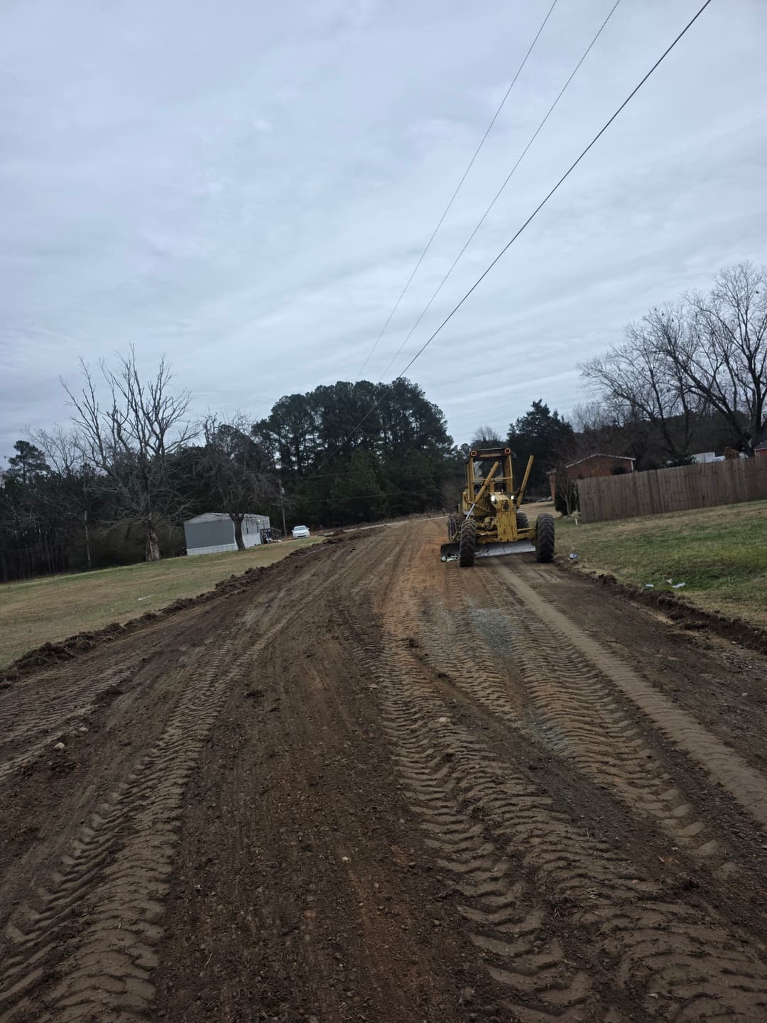 Grader machinery working on a dirt road with cloudy sky and trees in the background.