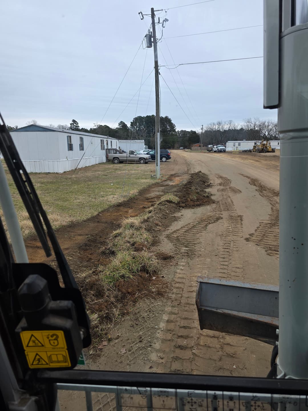 View from a construction vehicle showing a dirt road, power lines, and nearby buildings.