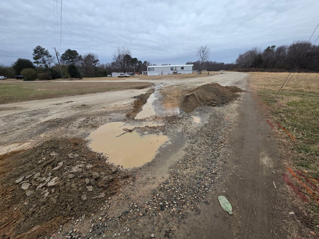 Pothole-filled gravel road with standing water and muddy excavations near a building.