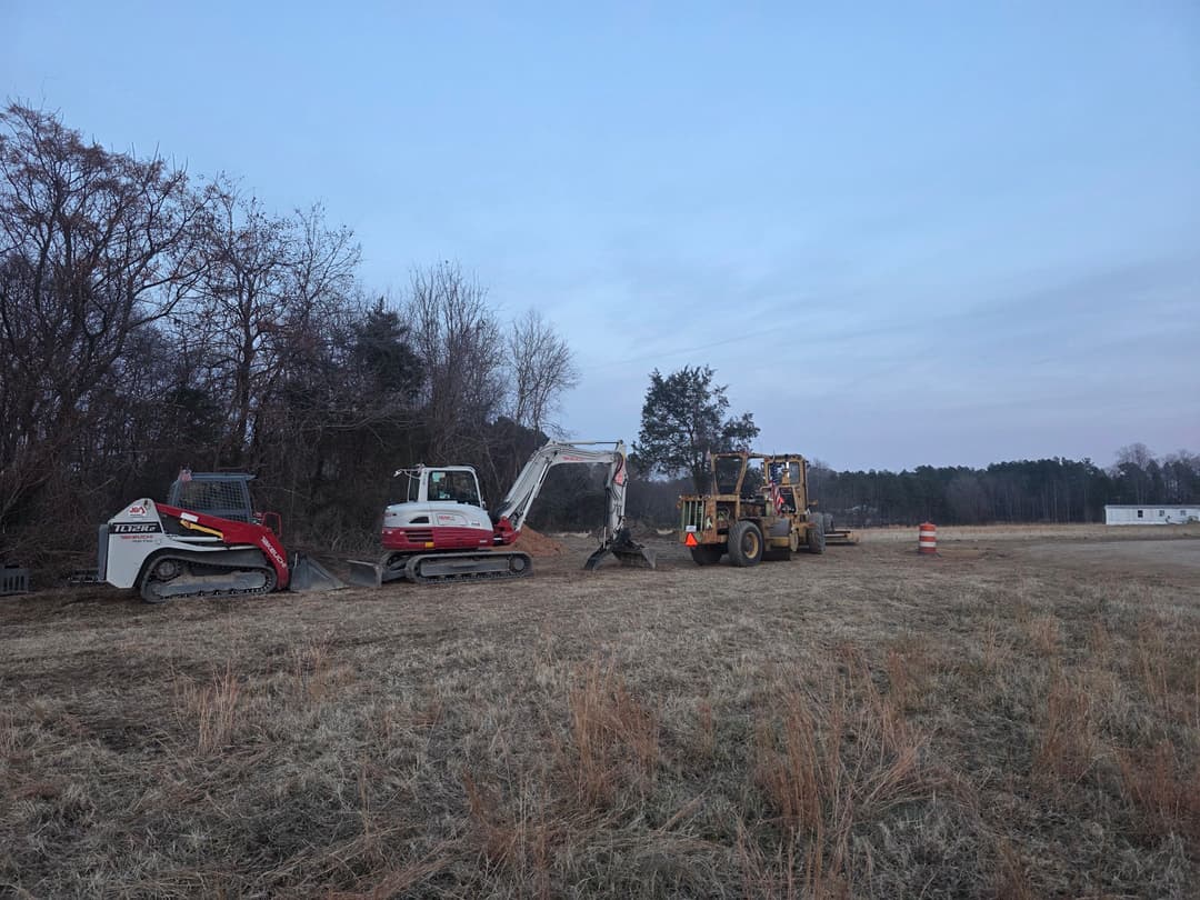 Construction workers using excavators on a rural site at dusk, surrounded by trees.