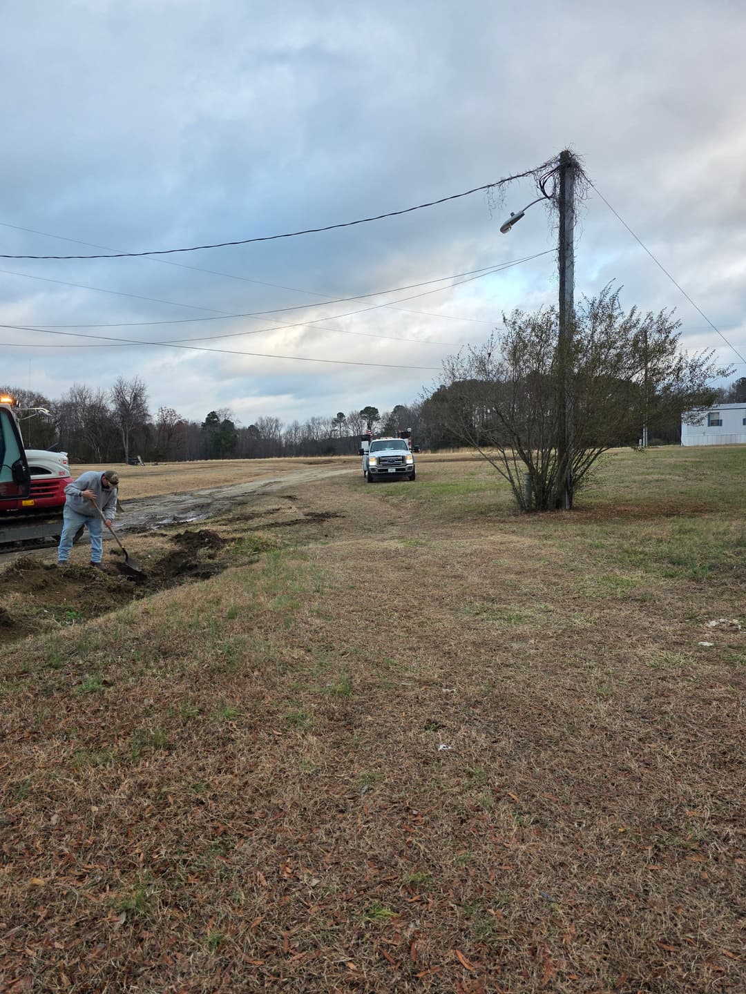 Construction worker digging near a truck in an open field under a cloudy sky.