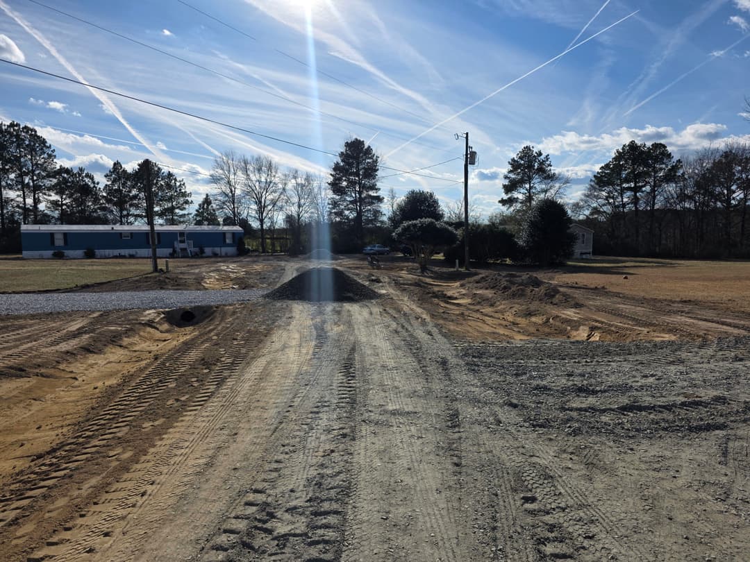 Construction site showing gravel roadwork, power lines, and scattered trees under a blue sky.