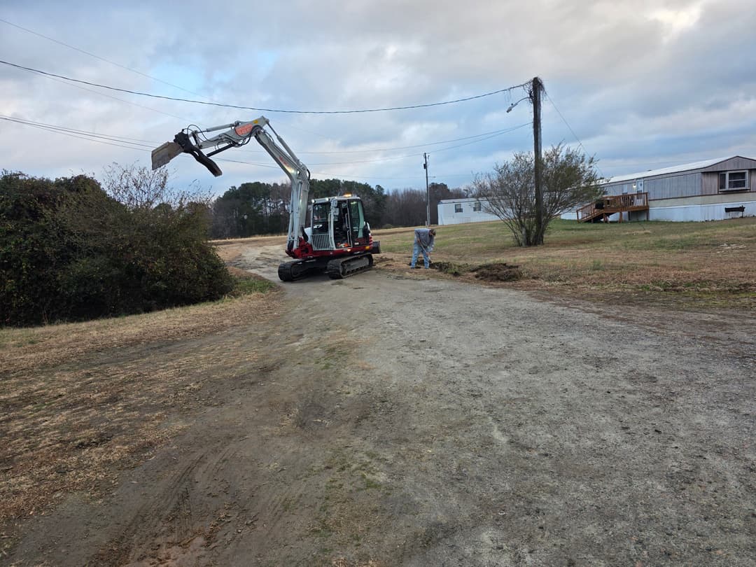 Excavator working on a dirt road with a worker in the background, cloudy sky above.