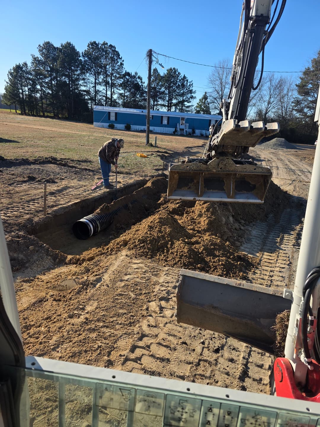 Excavator digging trench for drainage pipe installation in construction site. Worker present.