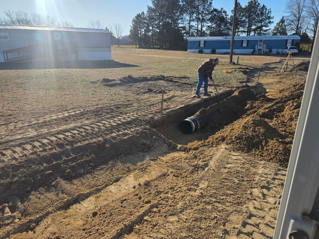 Worker installing a drainage pipe in a trench near mobile homes on a construction site.