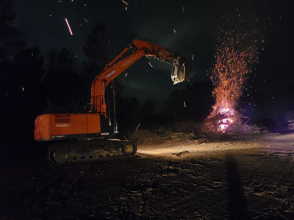 Excavator working at night near a bonfire with flying sparks in a wooded area.