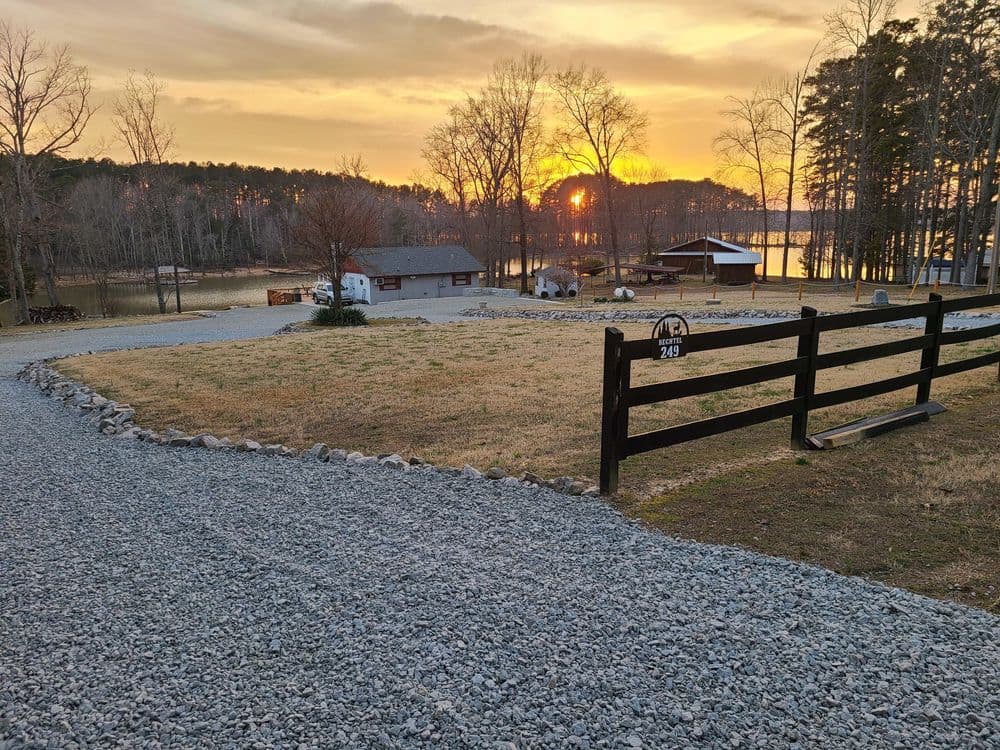Scenic sunset over a lake with gravel driveway and wooden fence at a peaceful countryside home.