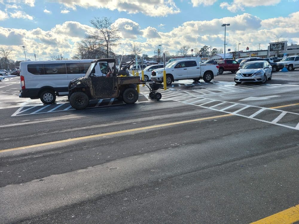 Utility vehicle painting parking lot lines under a cloudy sky, with cars parked nearby.