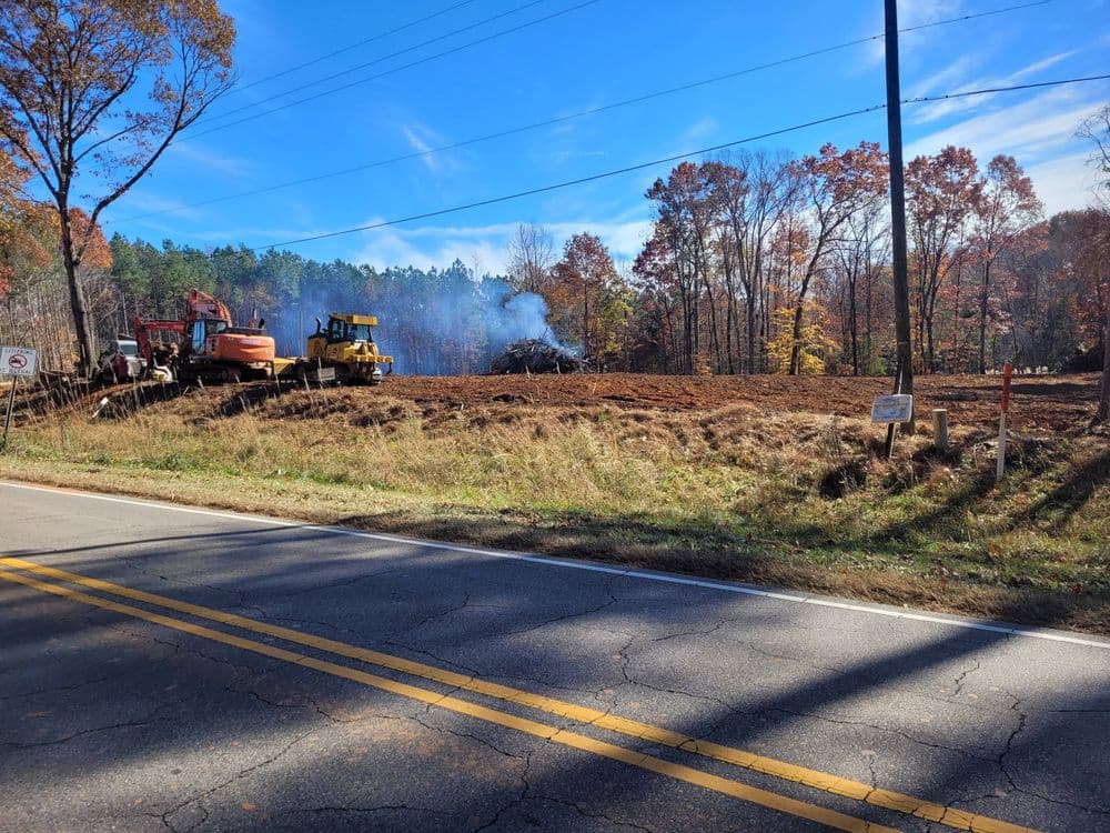 Construction equipment clearing land with smoke rising in a wooded area during autumn.